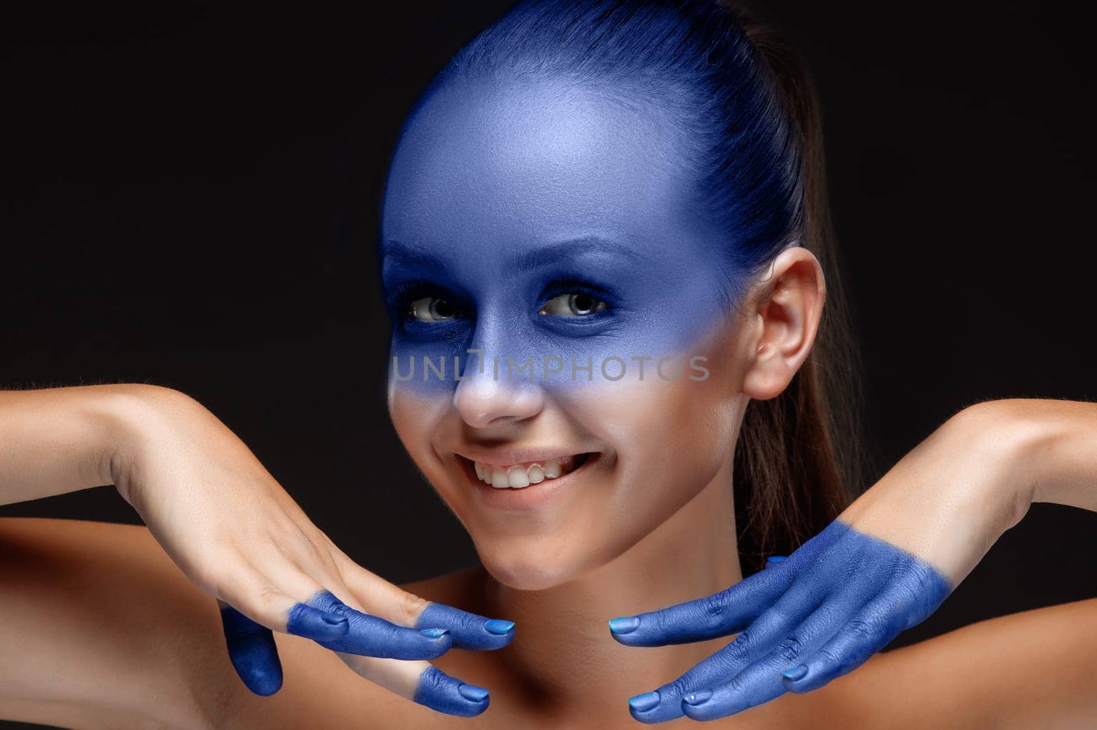 Portrait of a smiling young woman who is posing covered with blue paint in the studio on a black background. girl&#39 s hands around the face and painted blue