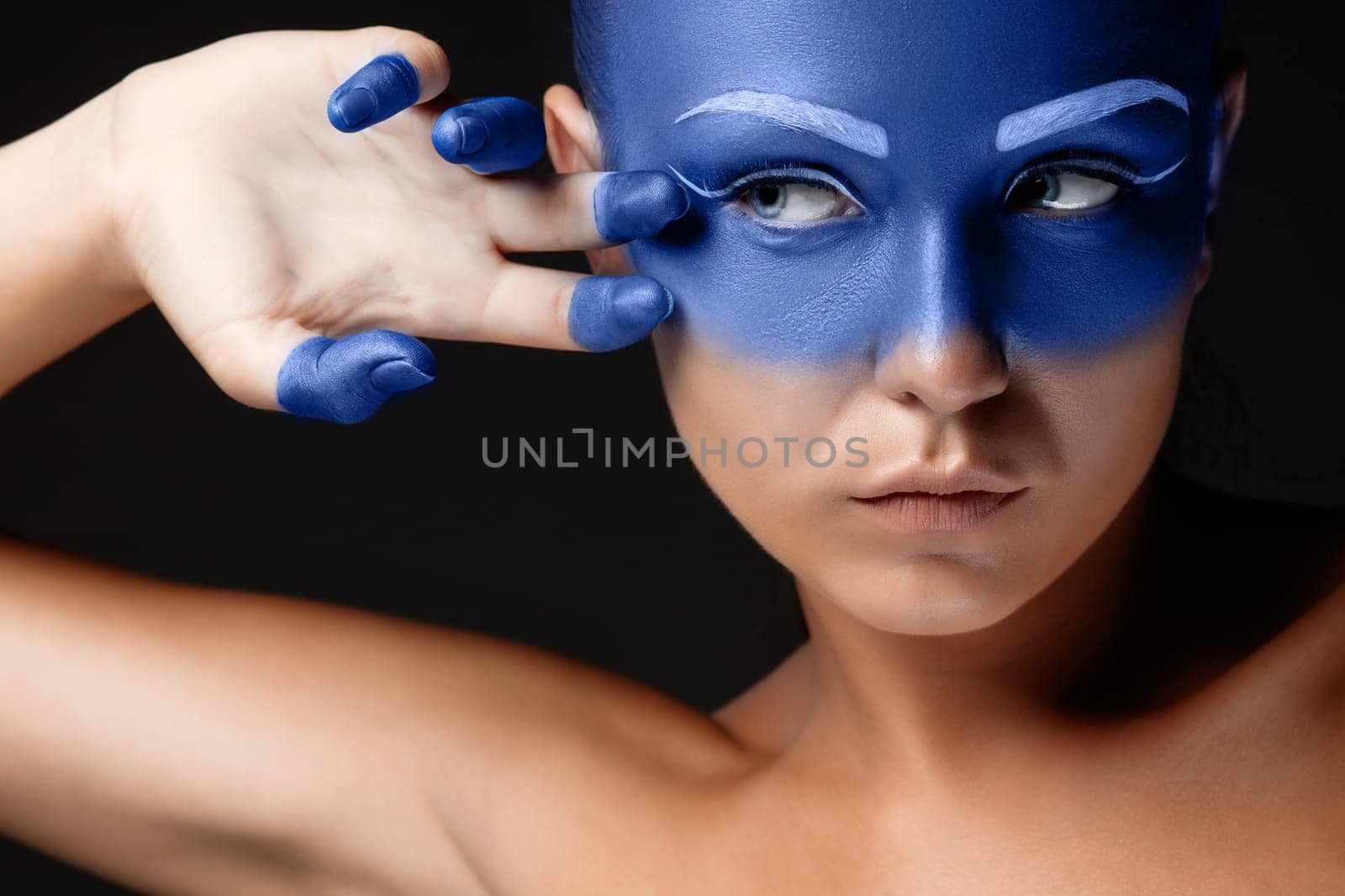 Portrait of a young woman who is posing covered with blue paint in the studio on a black background. girl&#39 s hand around the face and painted blue