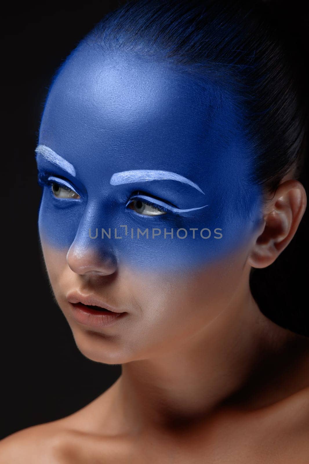 Portrait of a young woman who is posing covered with blue paint in the studio on a black background.