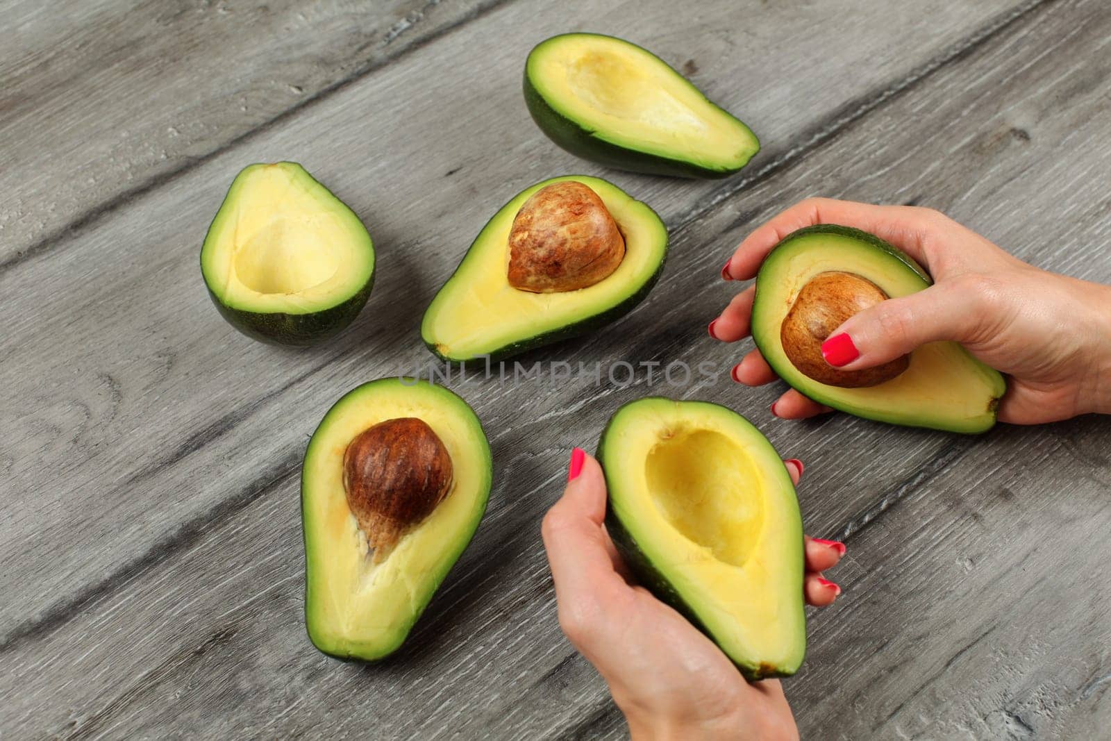 Avocado halves on gray desk, woman hands with red nails hold two of them.