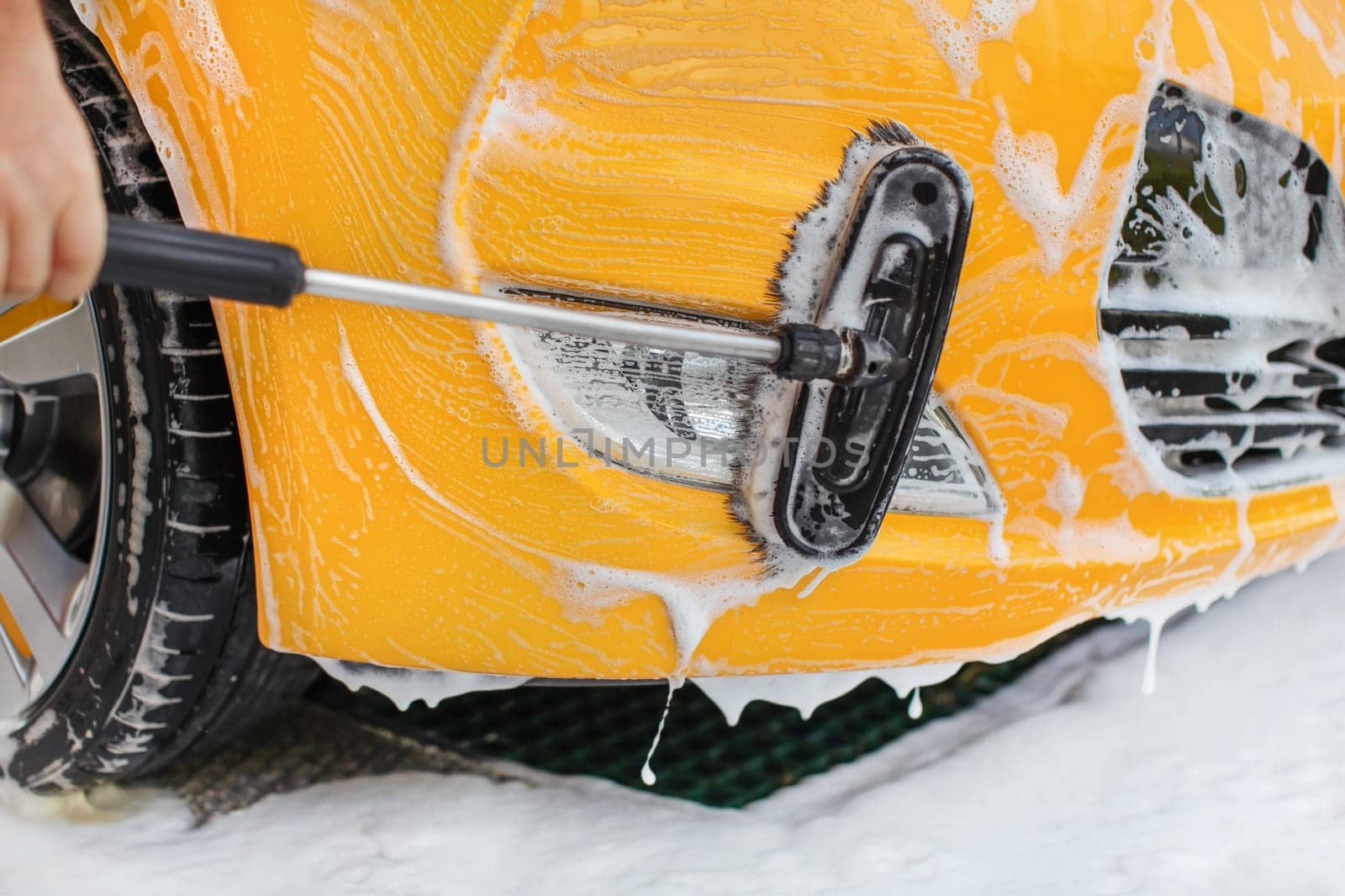 Front bumper and lights of yellow car washed in self serve carwash, brush  moving in white shampoo.