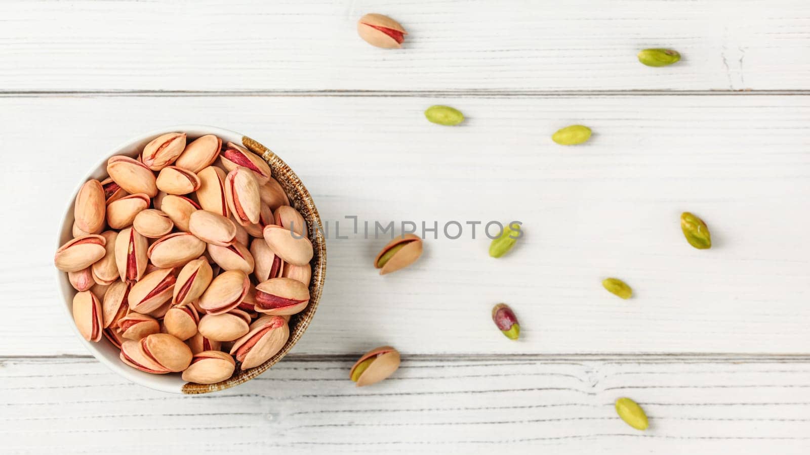 Tabletop view, small bowl with red Turkish pistachios, more nuts scattered on white board desk.