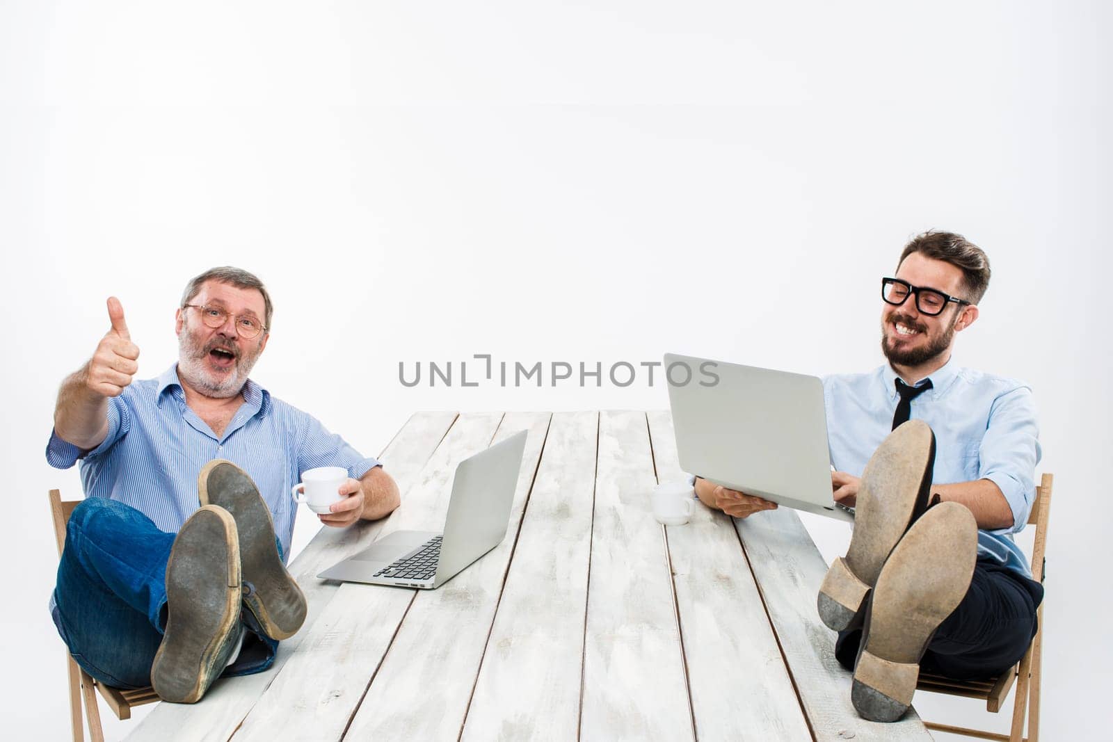 The two smiling businessmen with legs over table working on laptops on white background. Business in the American style