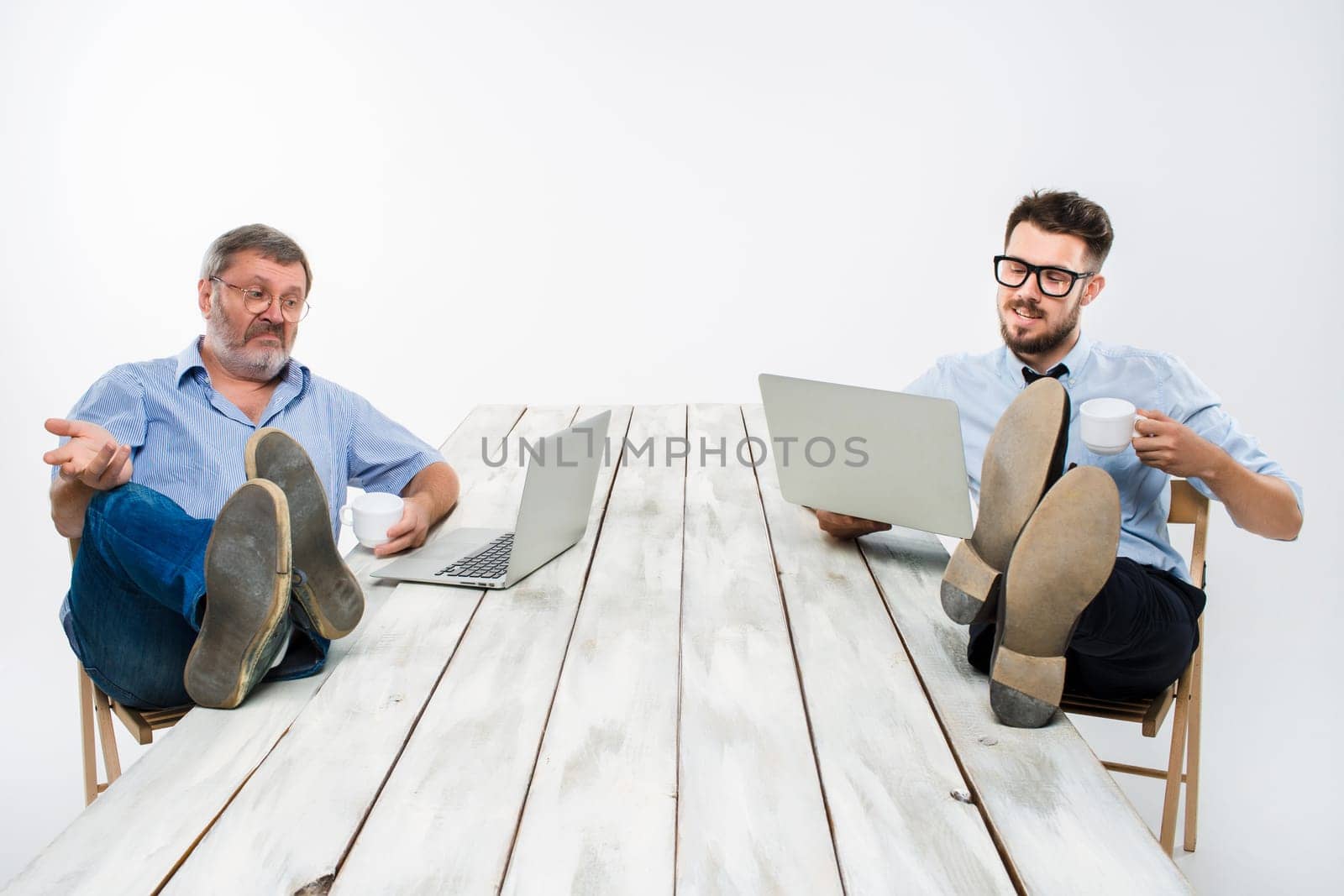 The two smiling businessmen with legs over table working on laptops on white background. Business in the American style