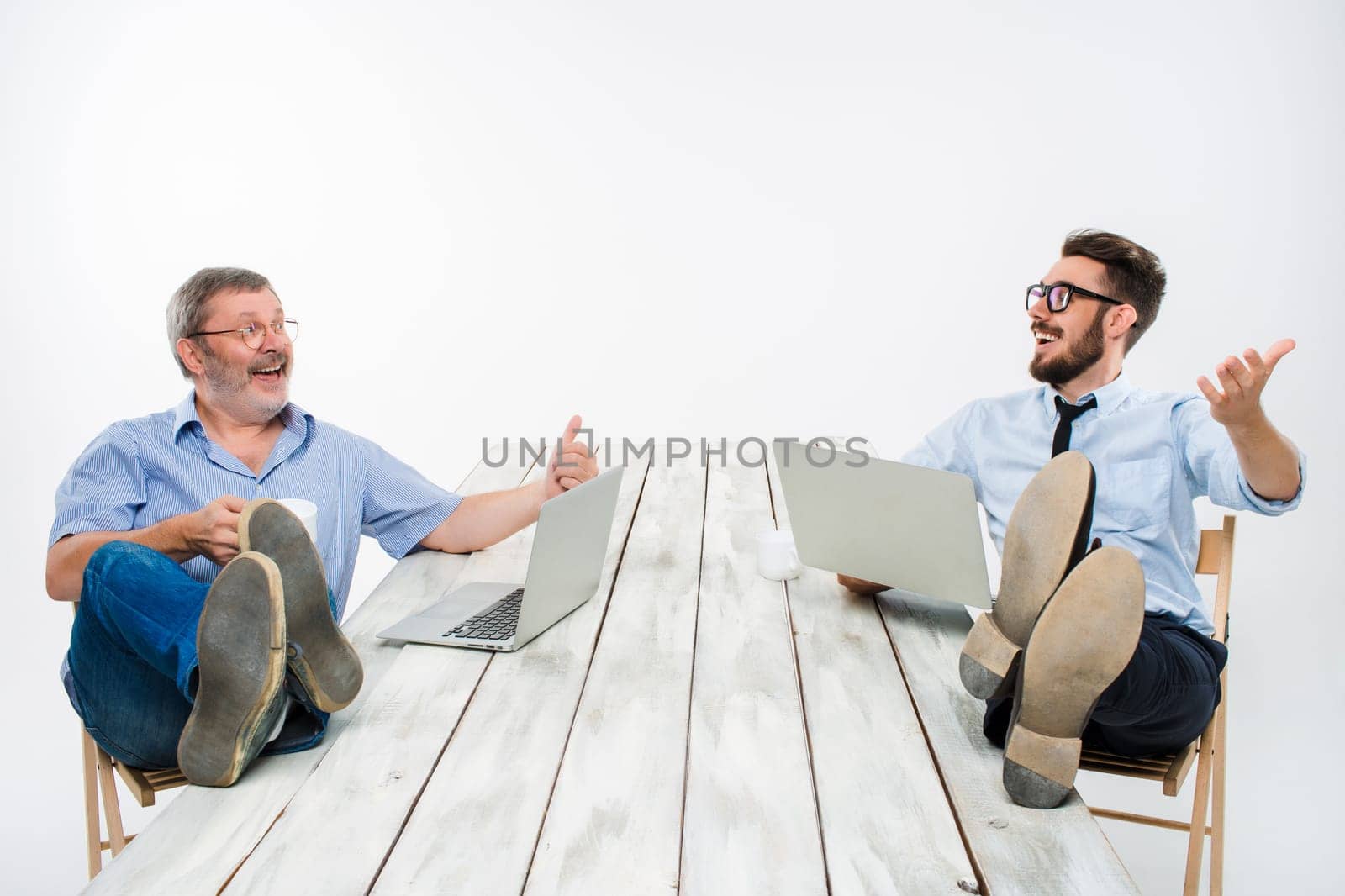 The two smiling businessmen with legs over table working on laptops on white background. Business in the American style