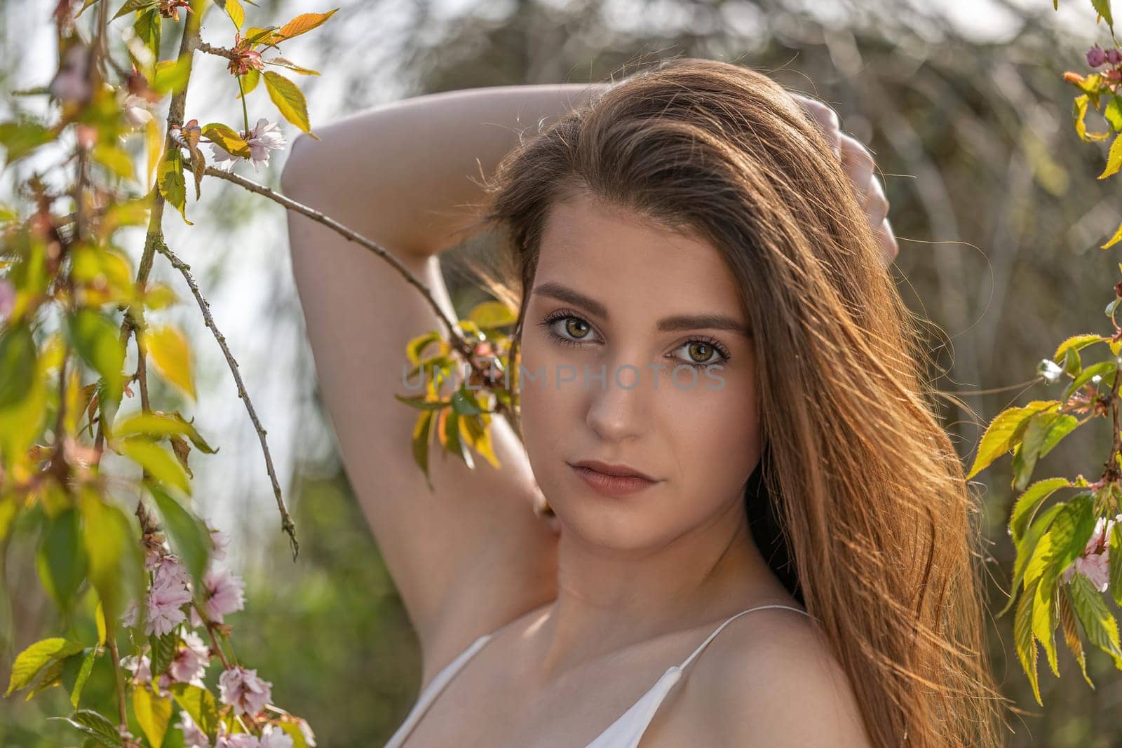 Cool brunette girl with hand in her long hair is posing on camera near sakura tree.. Horizontally.