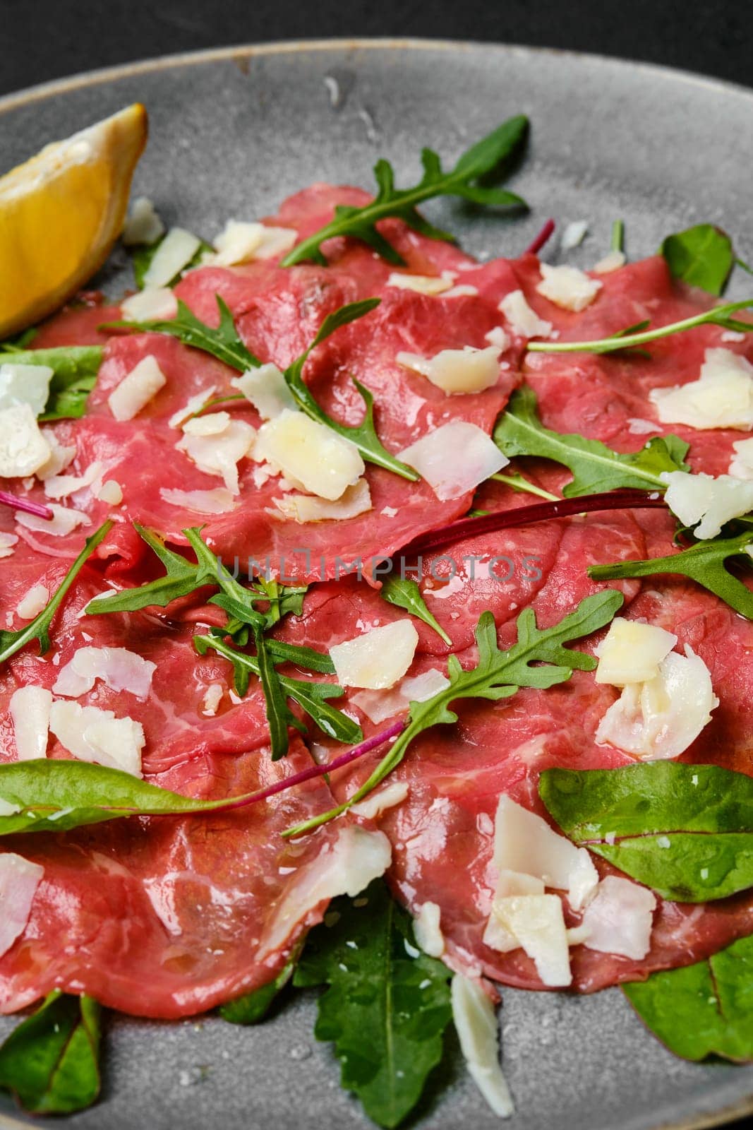 Marbled beef carpaccio with arugula and chard leaves, lemon and parmesan cheese on dark background, closeup