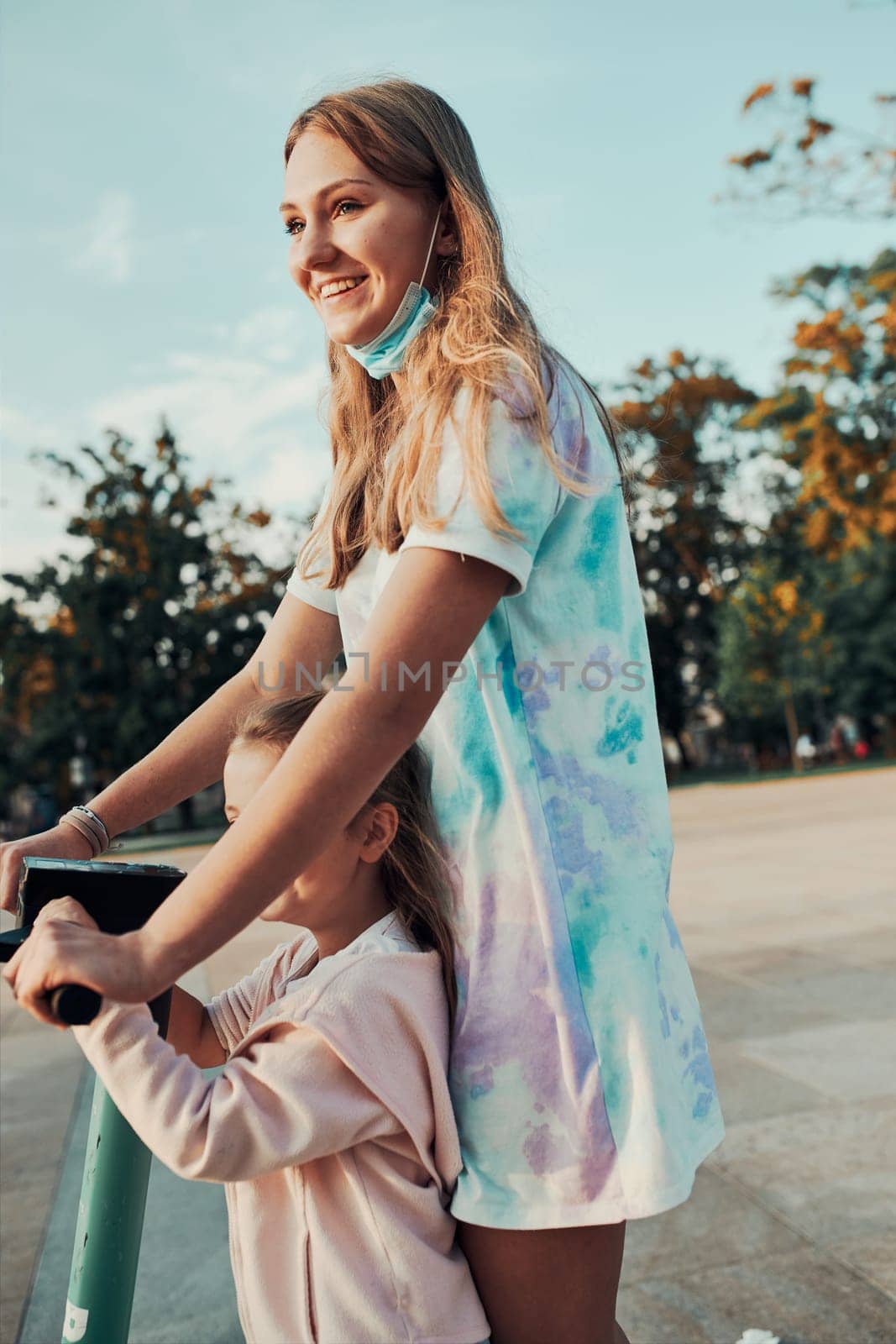 Teenager girl and her sister preschooler riding an electric scooter in the city center. Concept of happy family. Candid people, real moments, authentic situation