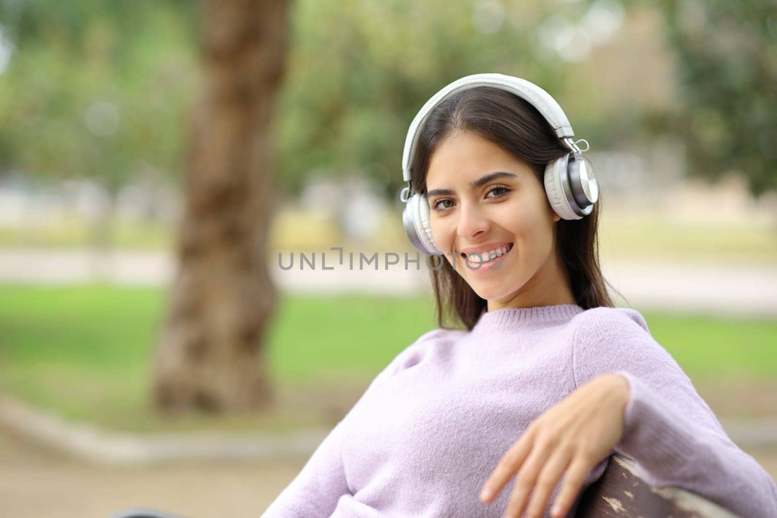 Happy woman listening to music looking at camera sitting on a bench in a park
