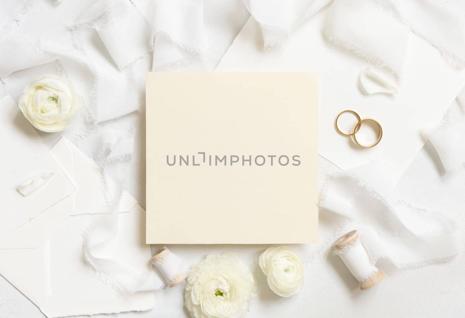 Blank card near cream roses, white silk ribbons and wedding rings top view, mockup. Romantic scene with square card and pastel flowers flat lay. Valentines, Spring or Mothers day concept