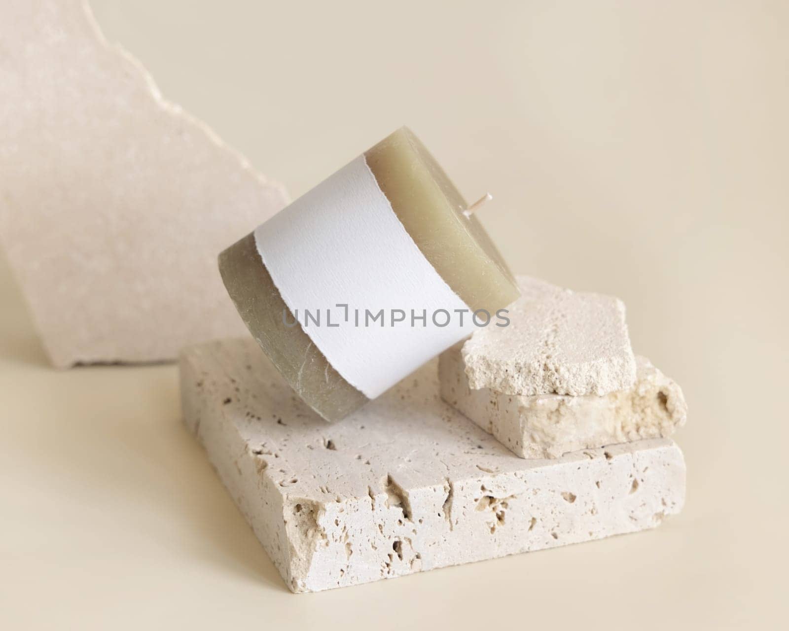 Candle with label on beige near cream travertine stones, Close up, copy space. Brand packaging mock up. Home decoration, minimal interior, Bohemian or scandinavian style