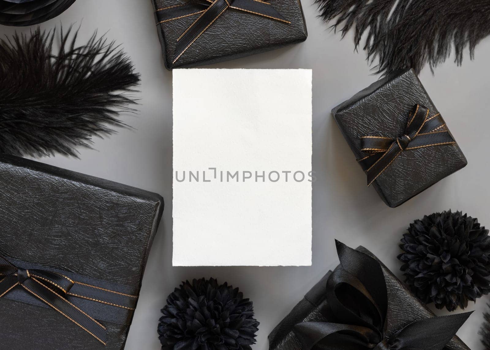 Blank card between Wrapped Gift Boxes, black paper flowers and feathers, mockup, copy space. Dark composition with vertical paper card for Birthday, Anniversary or black party present