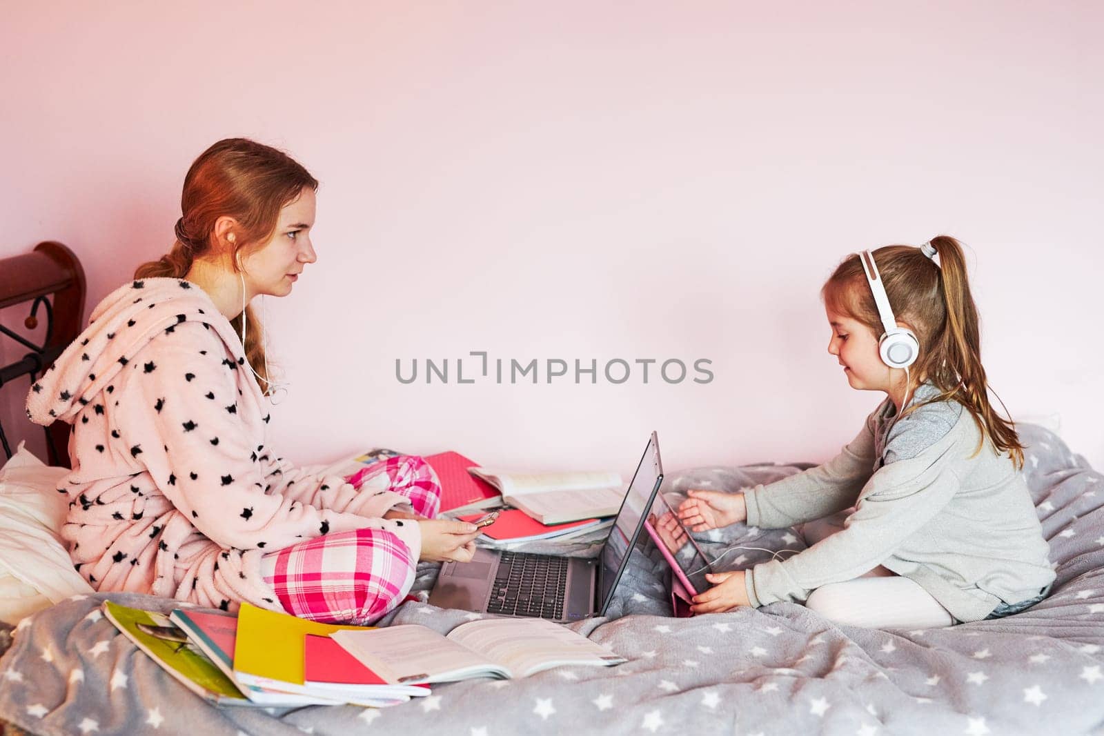 Student learning online, watching lesson remotely, listening to professor, talking with classmates on video call from home during quarantine. Young girl using laptop, headphones, books, manuals sitting in bed