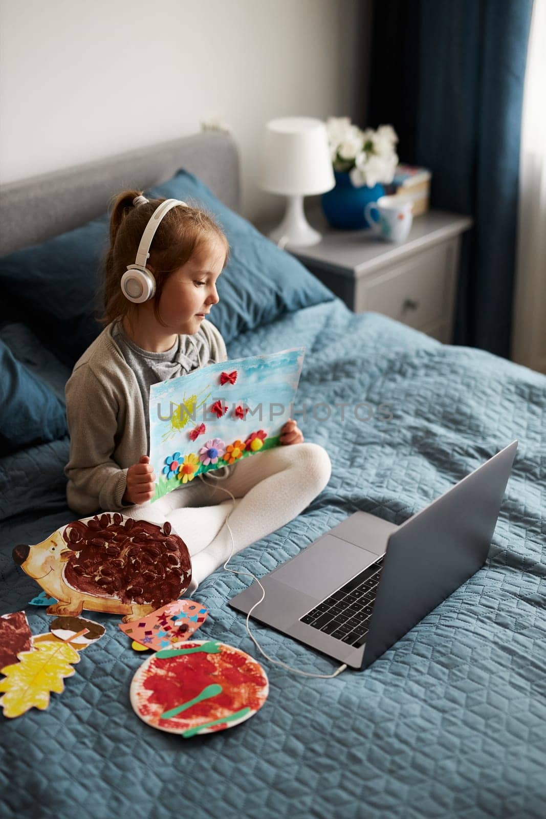 Little girl preschooler learning online showing her works drawings done at home. Child learning watching lesson remotely, talking with tutoress on video call from home during quarantine. Kid using laptop, headphones sitting on bed