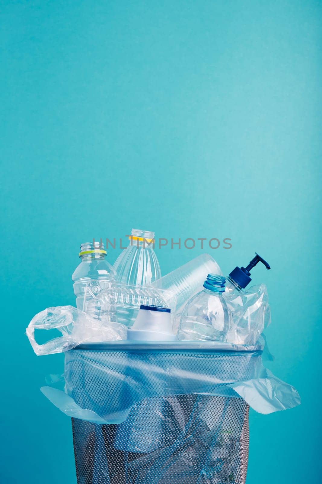 Heap of plastic bottles, cups, bags collected to recycling in a metal bin. Concept of plastic pollution and too many plastic waste. Trash with used plastic packagings over blue background with copy space at the top