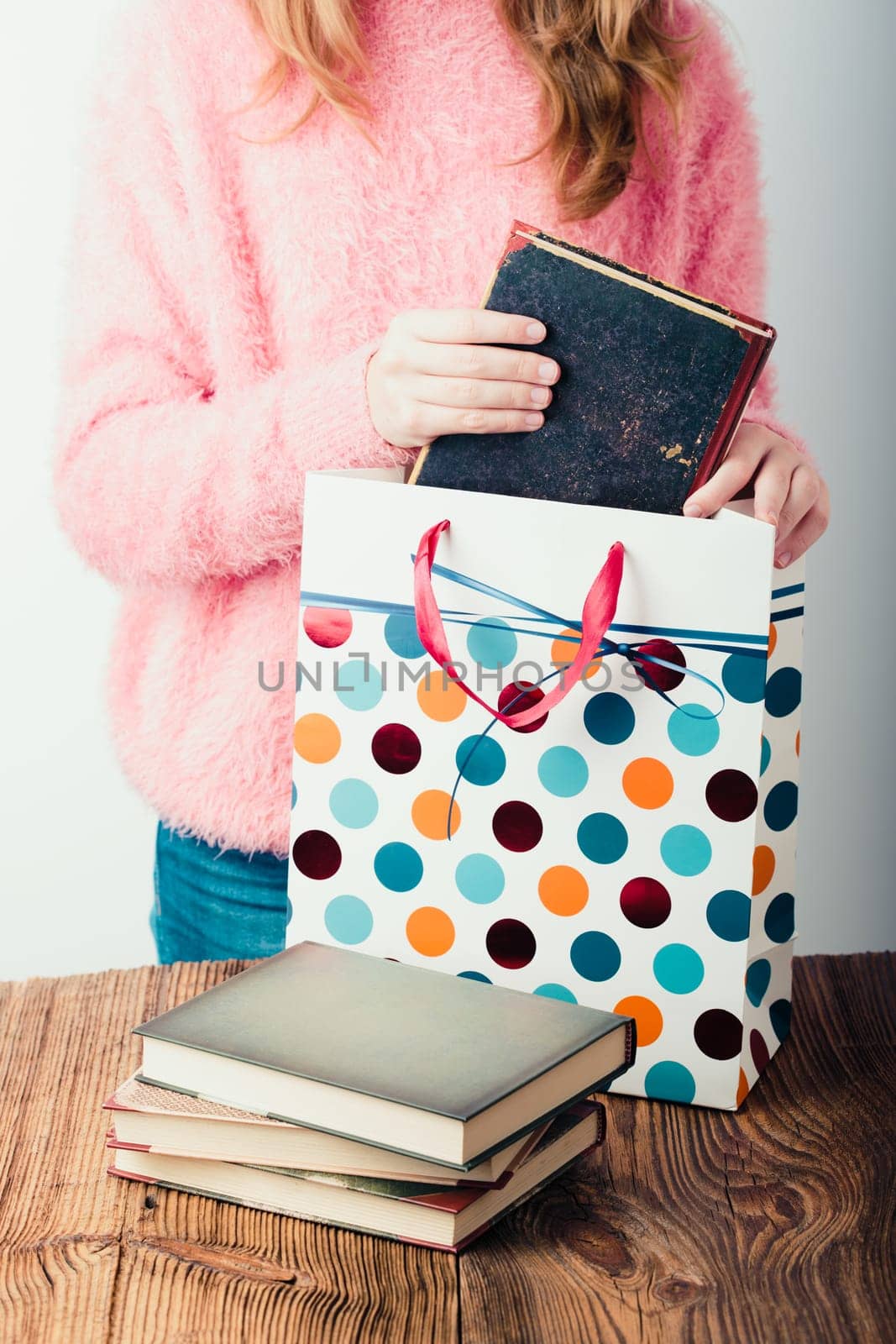 Young girl putting the books into paper bag in bookstore. A few books on a wooden table. Teenager girl wearing pink sweater and blue jeans. Vertical photo