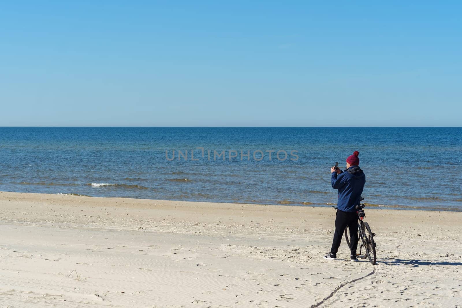 A man in warm clothes and a hat takes a photo on his phone standing near his bicycle, traveling along the Baltic Sea on the Curonian Spit, Neringa, Lithuania.