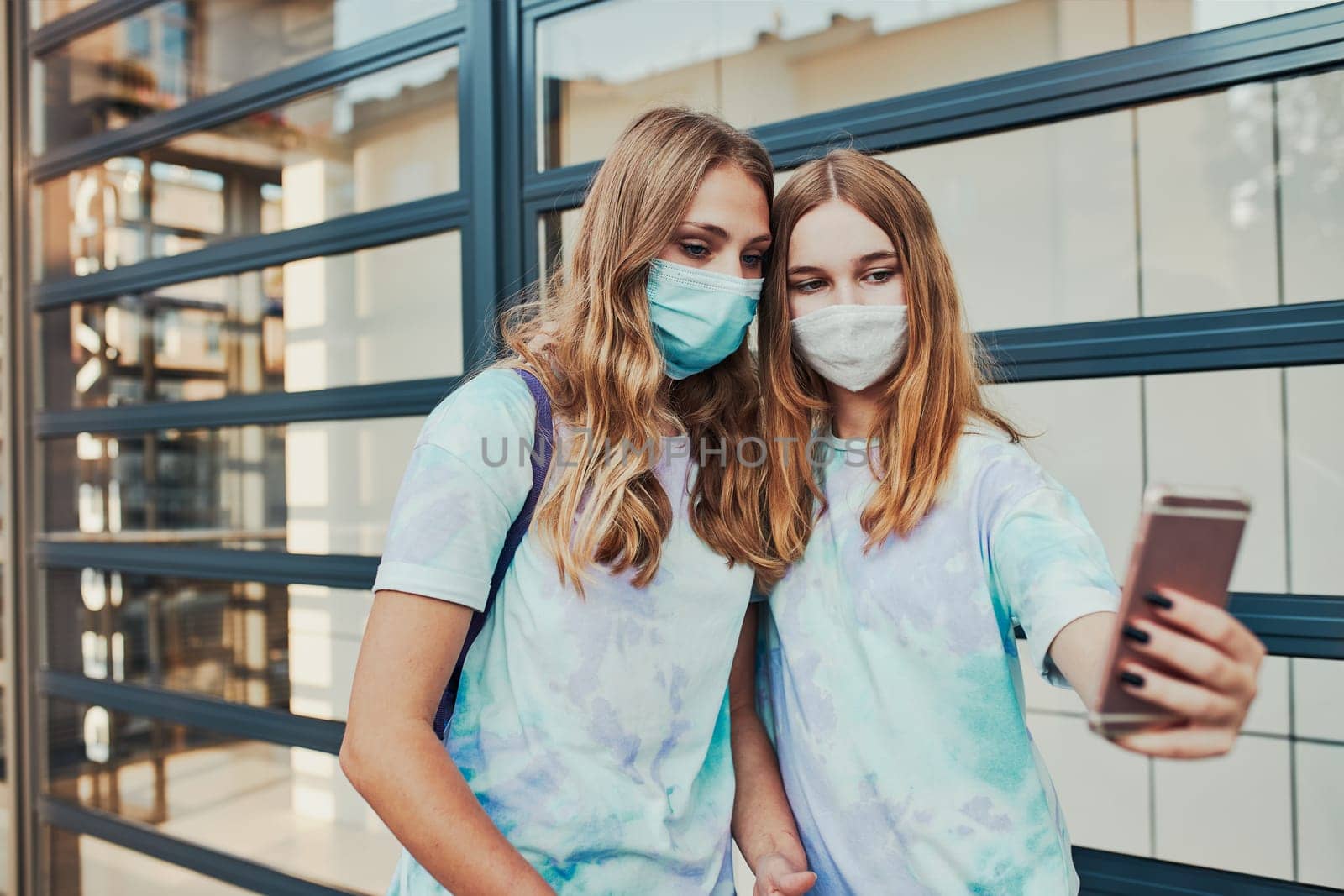 Young women girls taking selfie photos using smartphone standing at store front downtown. Girls wearing face masks to avoid virus infection and to prevent the spread of disease in time of coronavirus