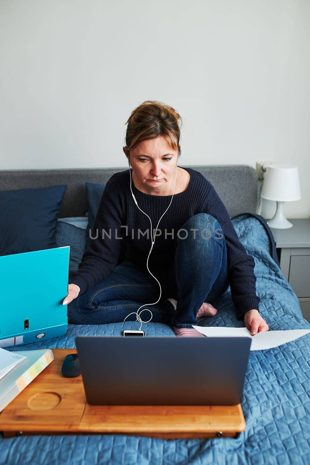 Woman working doing her job remotely during video chat call stream online course webinar on laptop from home. Woman sitting on bed in front of computer looking at screen using headphones and smartphone