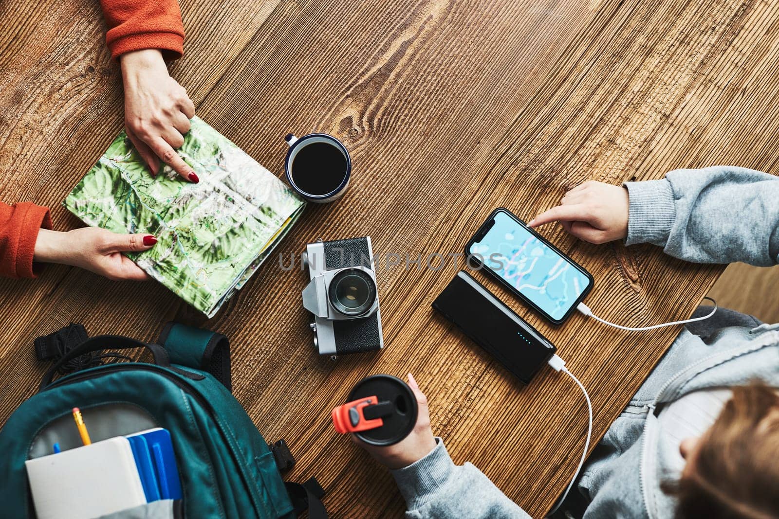 Family planning vacation trip. Girl searching travel destination and routes using navigation map on mobile phone. Woman pointing on paper map. Charging smartphone with power bank. Using technology while travelling. People sitting at wooden table with camera and backpack during journey. Travel plan concept