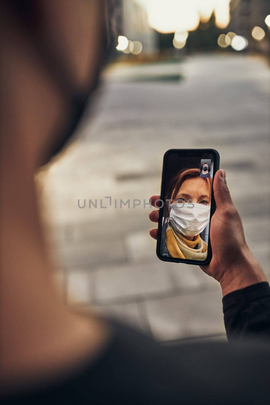 Young man having video call with her mother standing outdoors in the street in the evening wearing the face mask to avoid virus infection and to prevent the spread of disease in time of coronavirus