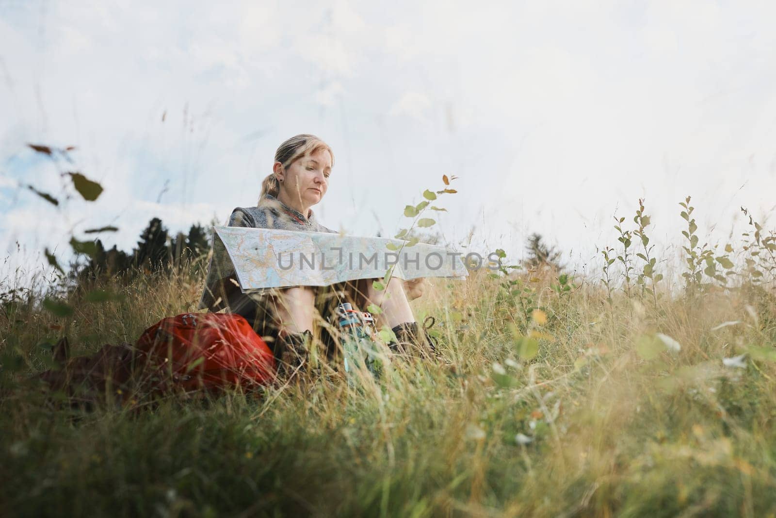 Woman with backpack having break during trip in mountains looking at map sitting on grass on summer vacation day