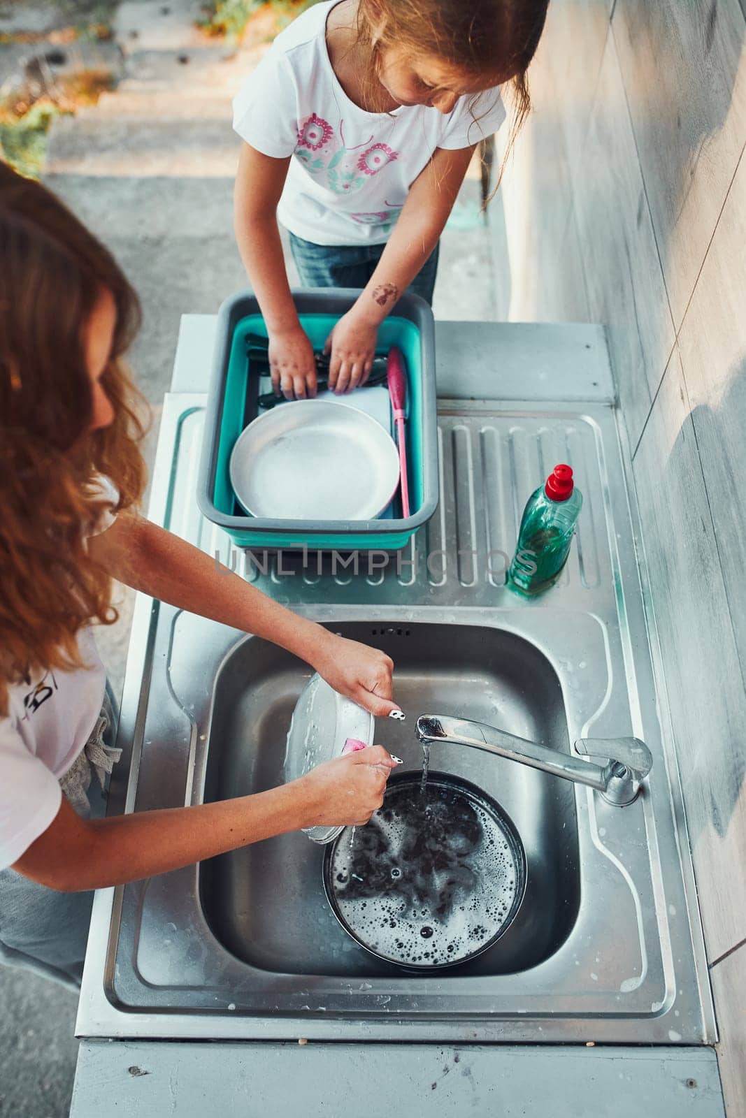 Teenager girl washing up the dishes pots and plates with help her younger sister in the outdoor kitchen during vacations on camping. Camp life. Sisters spending time and working together