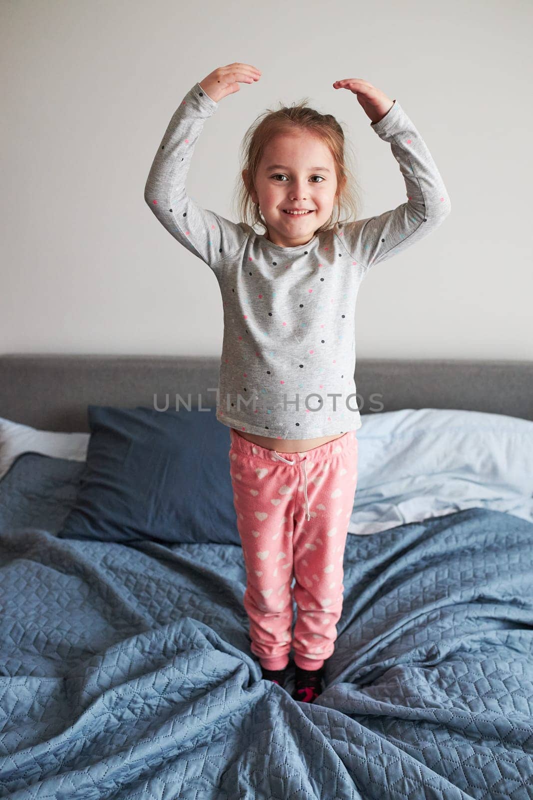 Little happy playful kid girl having fun making silly funny faces jumping on bed in bedroom in the morning at home