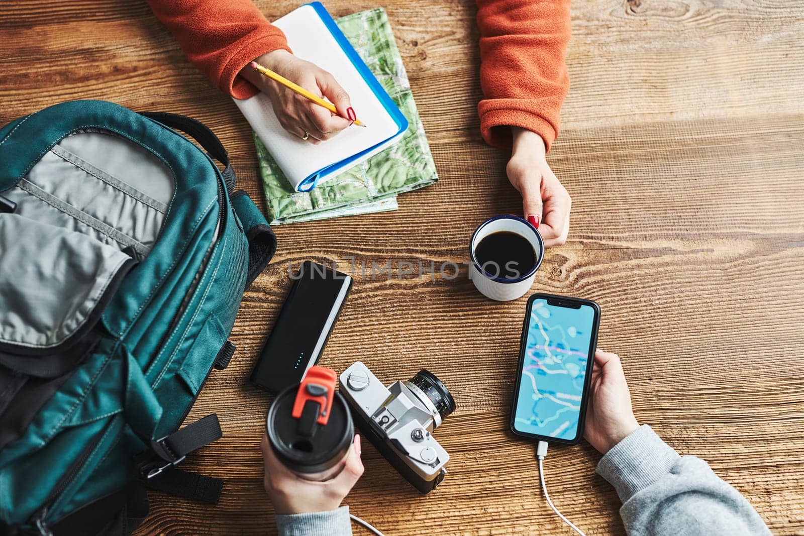 Family planning vacation trip. Girl searching travel destination and routes using navigation map on mobile phone. Woman writing journal and making notes. Charging smartphone with power bank. Using technology while travelling. People sitting at wooden table with camera and backpack during journey. Travel plan concept