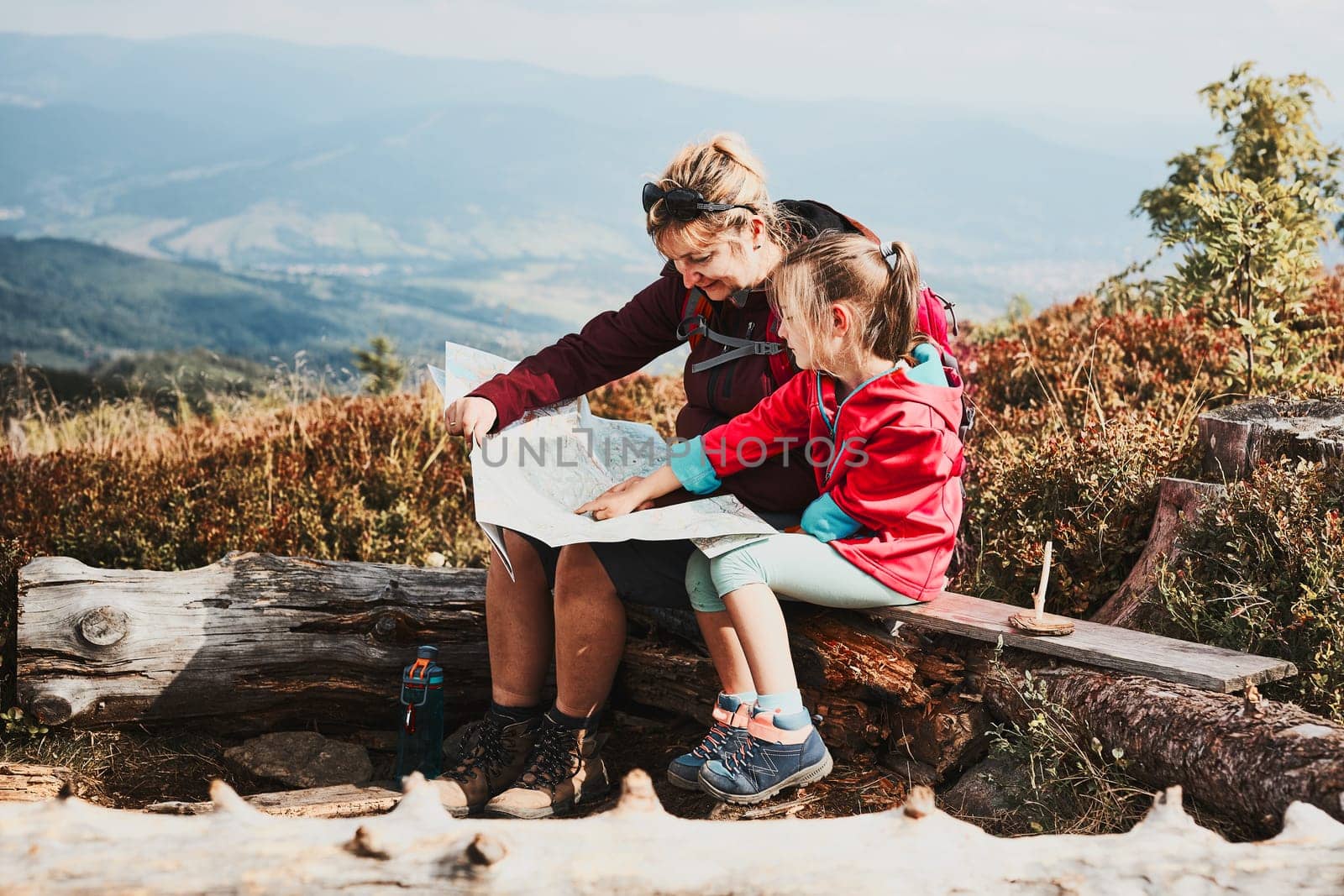 Family trip in mountains. Mother and her little daughter examining a map, sitting on stump, taking break during summer trip