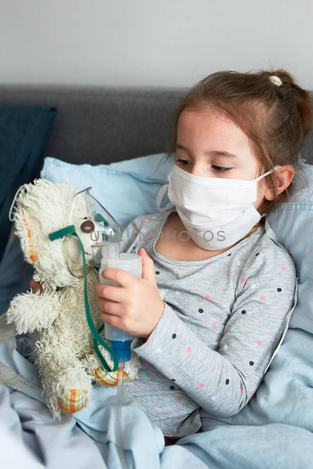 Sick child recovering in bed. Little girl playing by applying medical inhalation treatment with nebuliser to her teddy bear