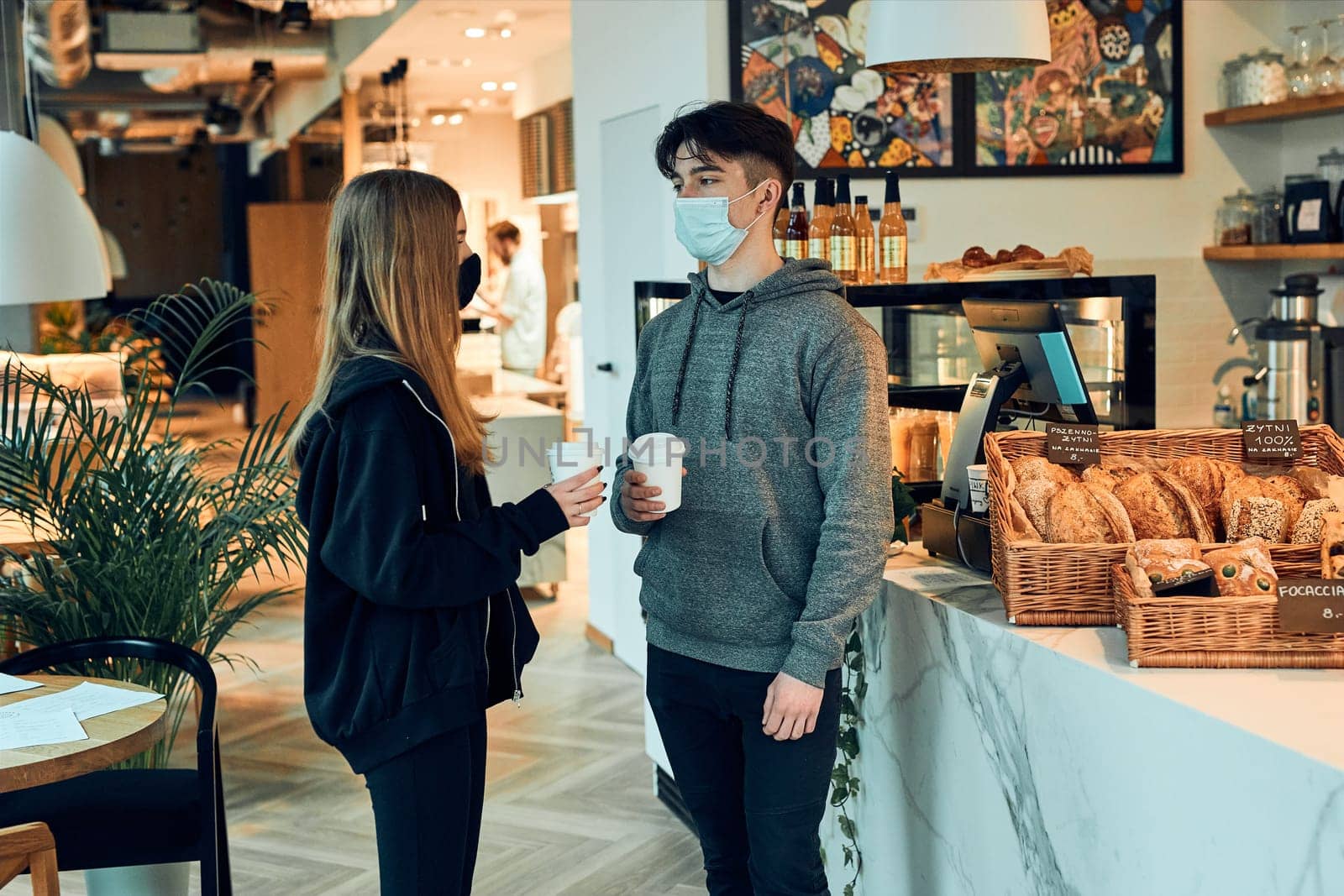Friends having a chat, holding coffee cups, standing in a coffee shop and waiting for the orders. People wearing the face masks to avoid virus infection and to prevent the spread of disease in time of coronavirus