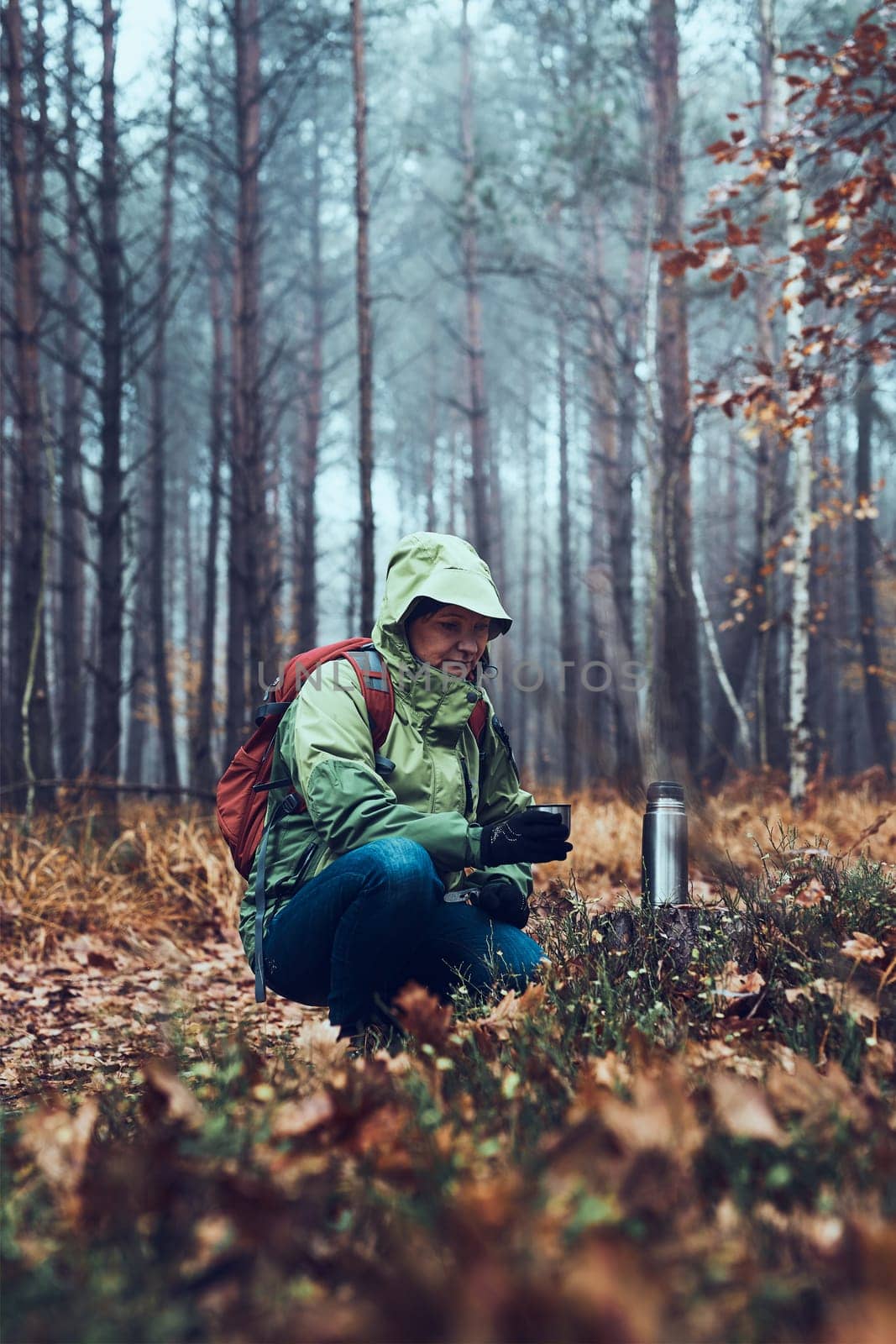 Woman with backpack having break during autumn trip drinking a hot drink from thermos flask on autumn cold day. Active middle age woman wandering around a forest actively spending time