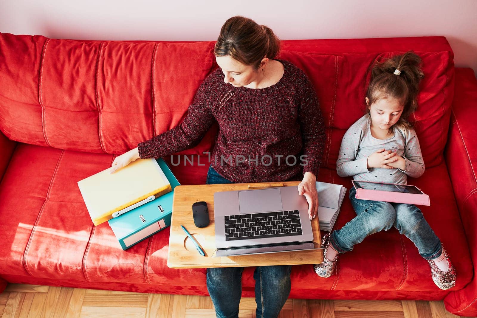 Woman mother working doing her job remotely during video chat call stream online course webinar on laptop from home during COVID-19 quarantine while her daughter watching video on tablet. Woman sitting on bed in front of computer looking at screen