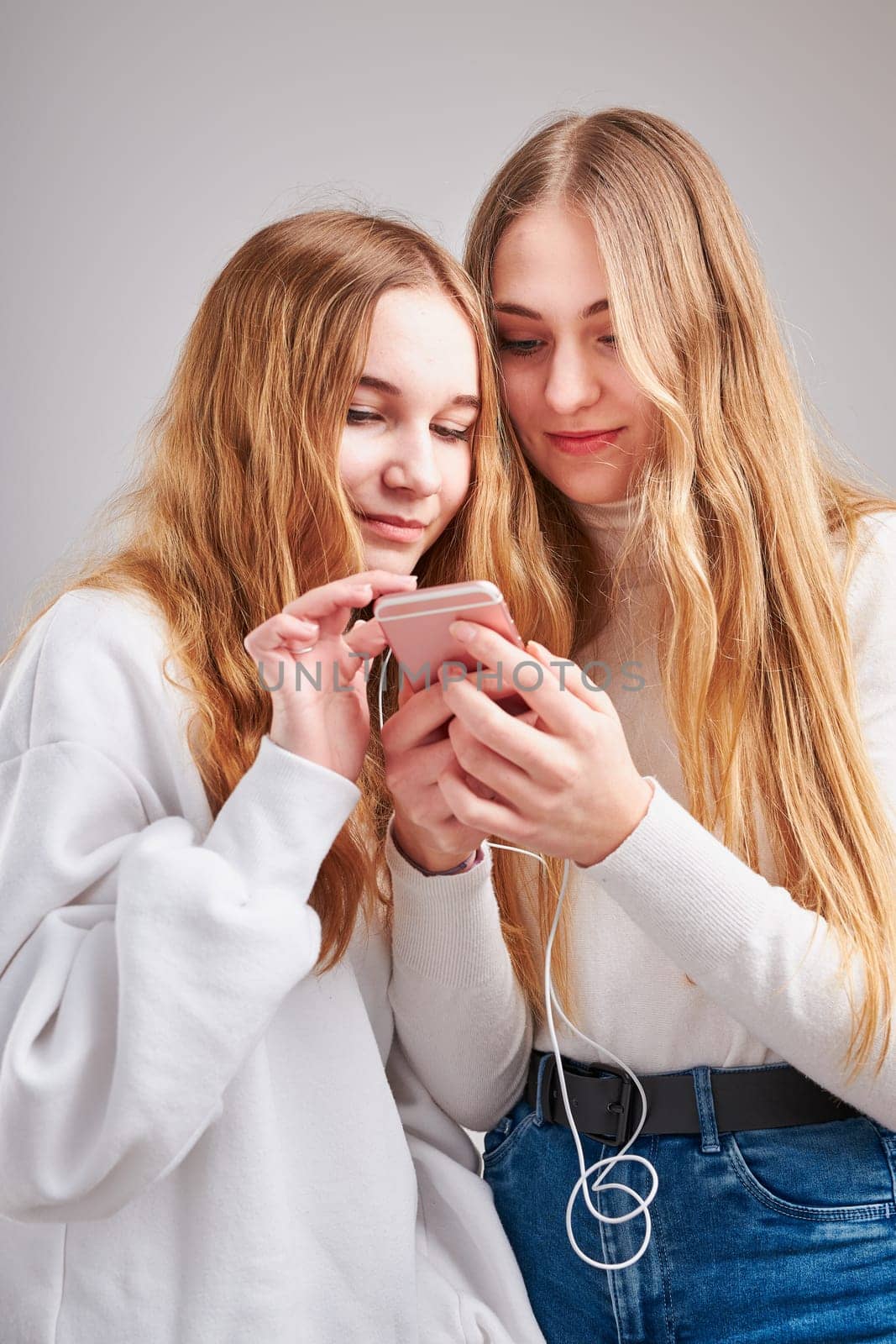 Young women girls listening to music together streaming content having fun watching video enjoying video chat talking with friends making gestures faces using smartphone earphones headphones standing over plain grey background
