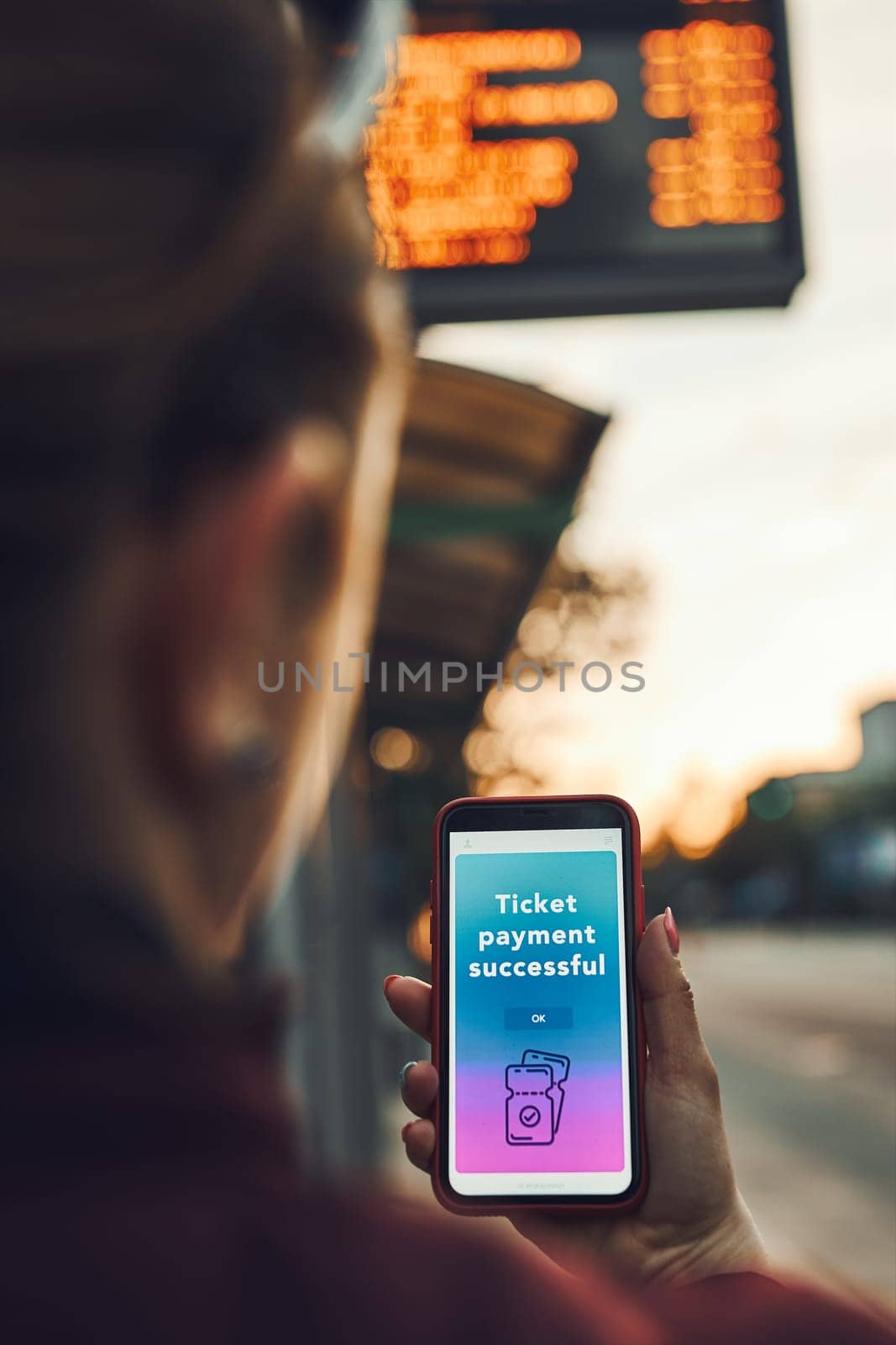 Woman buying ticket online near bus stop before ride. Person paying for ticket using mobile payment app. Using technology on the go. Order confirmation on mobile screen. Top up account in app. Bus routes schedule in background
