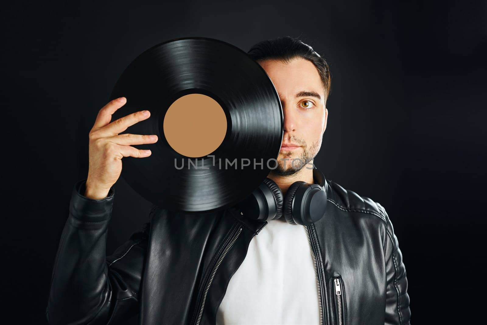 Man holding black vinyl record over face. Vintage music style. Rock style. Male wearing black jacket holding turntable disc standing on dark background. Retro music. Classic audio