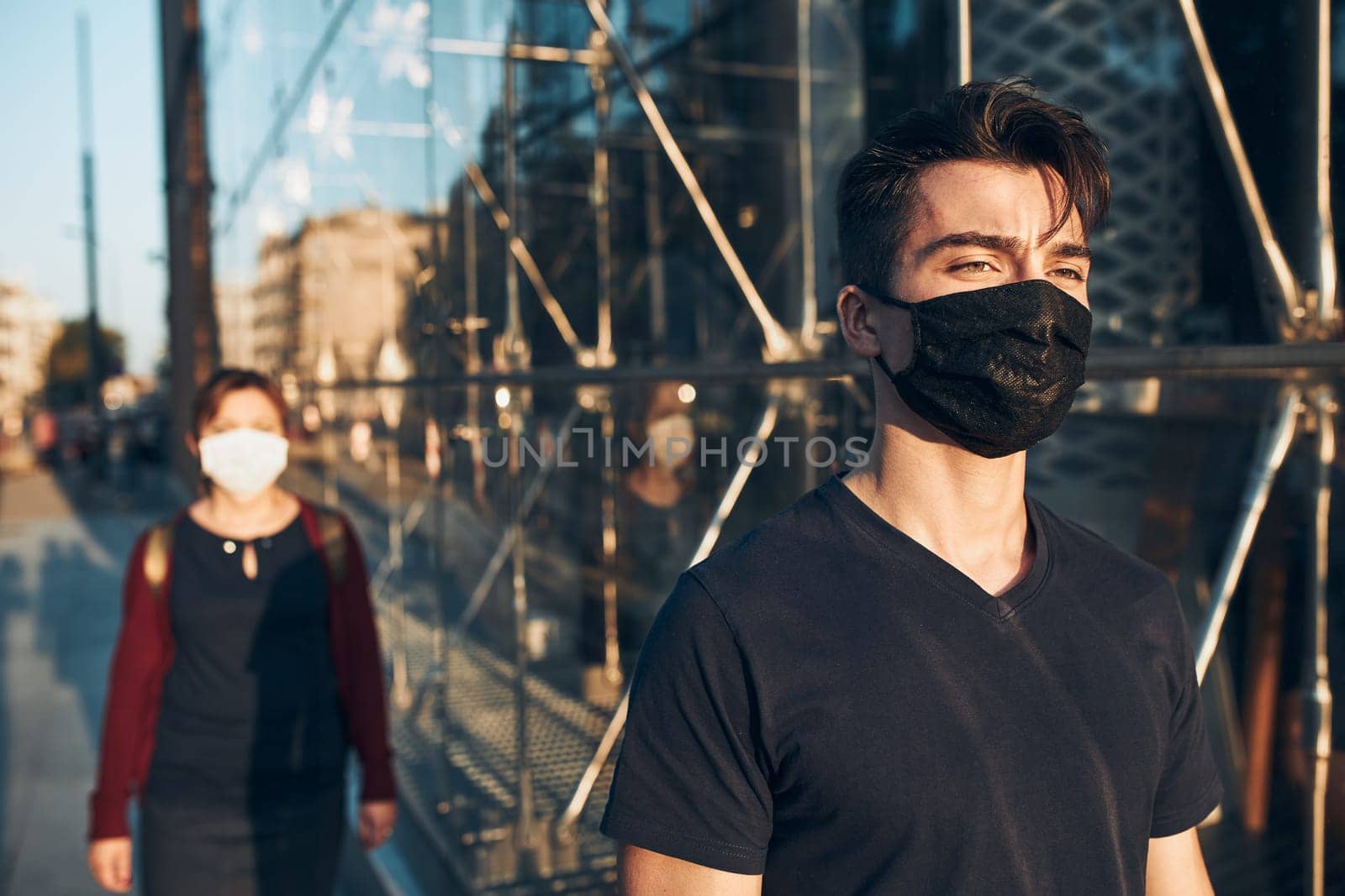 Young man walking in the city center along glass store front in the evening, looking away, wearing the face mask to avoid virus infection and to prevent the spread of disease in time of coronavirus