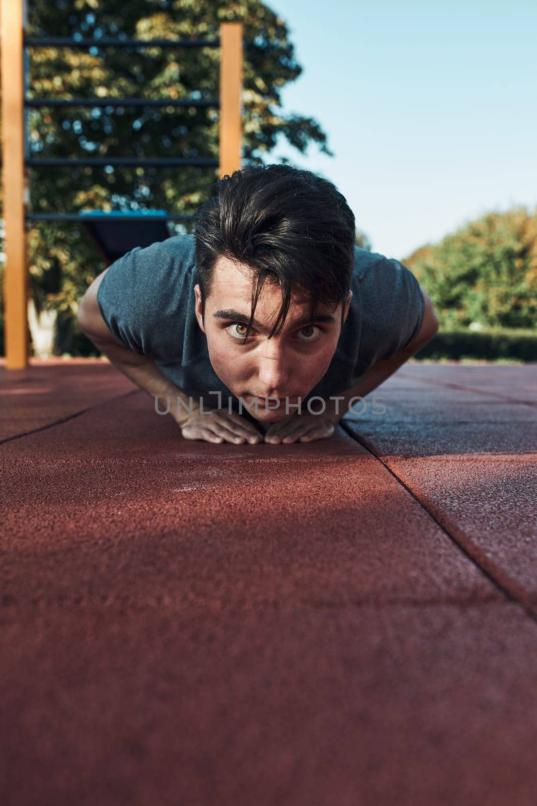 Young man doing push-ups on a red rubber ground during his workout in a modern calisthenics street workout park. Man wearing sportswear looking at camera