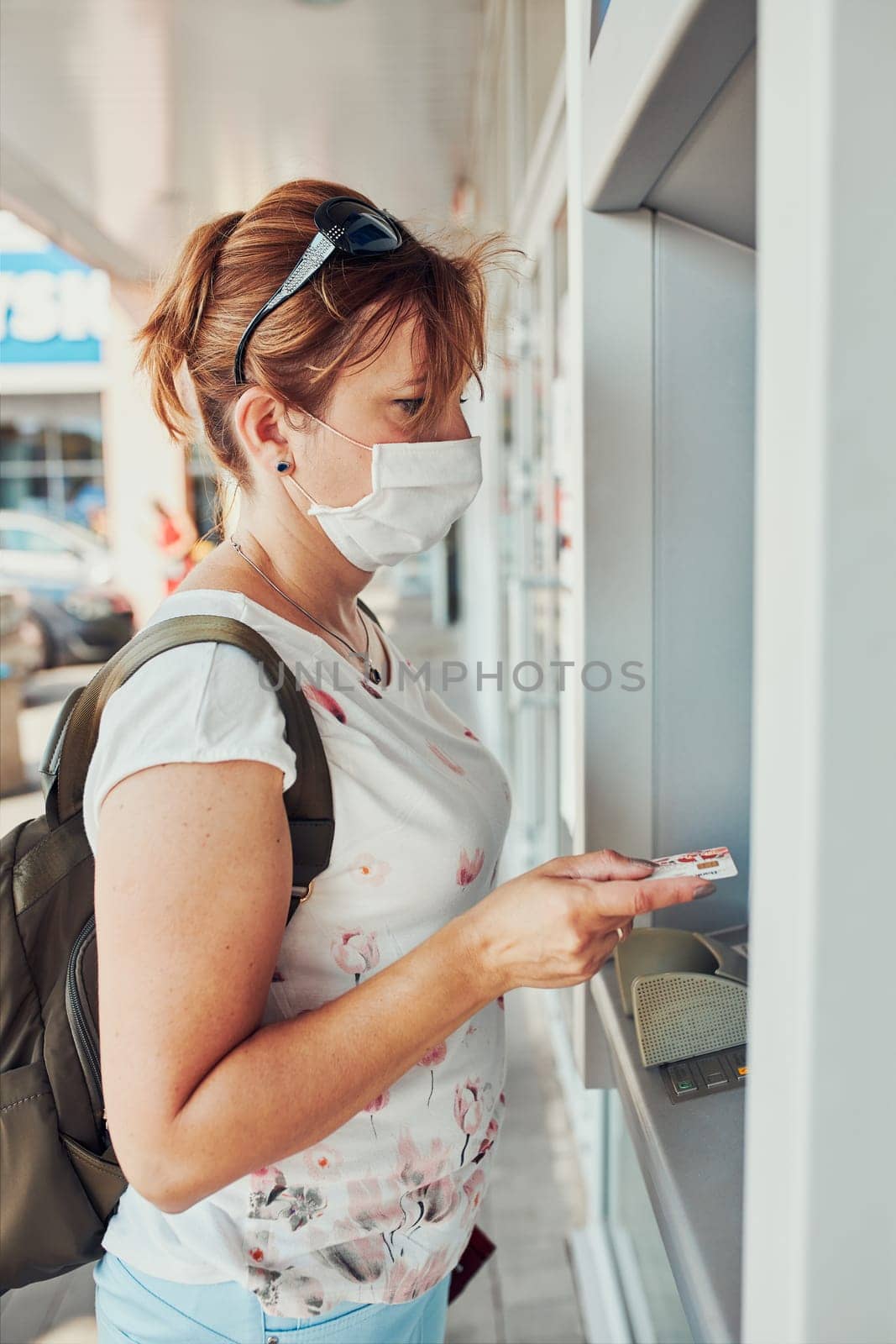 Woman withdrawing money from atm machine using debit or credit card standing outdoors in the street, wearing face mask to cover mouth and nose during pandemic coronavirus outbreak