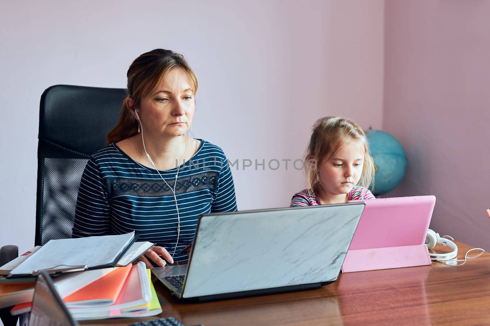 Woman mother working doing her job remotely during video chat call stream online course webinar on laptop from home while her daughter watching video on tablet. Woman sitting at desk in front of computer looking at screen