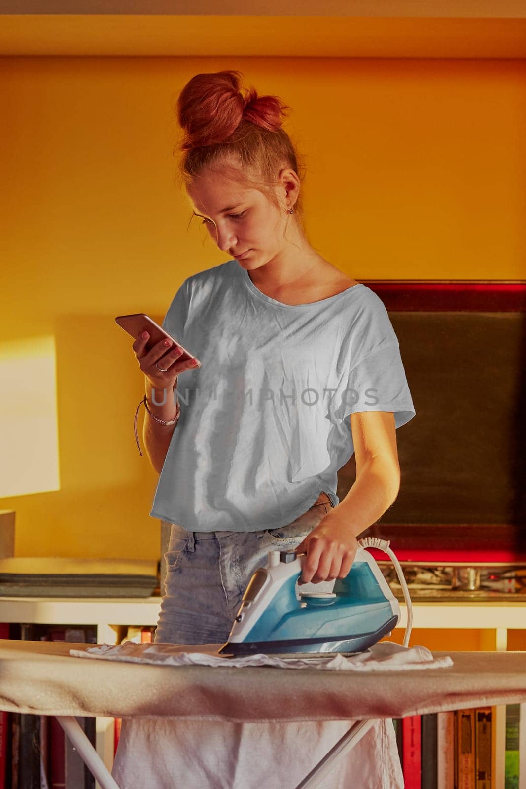 Young woman is ironing her clothes and talking on a smartphone simultaneously, standing by a ironing board in a room at home. Routine housekeeping task at home. Candid people, real moments, authentic situations