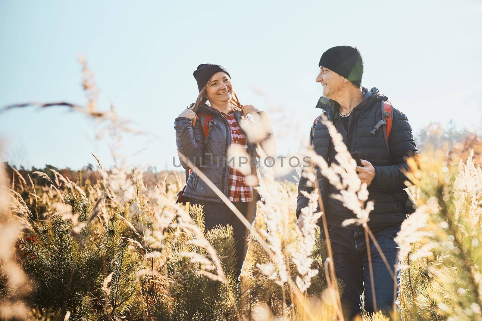 Couple having fun while vacation trip. Hikers with backpacks on way to mountains. People exploring nature walking through tall grass along path in meadow on sunny day. Active leisure time close to nature