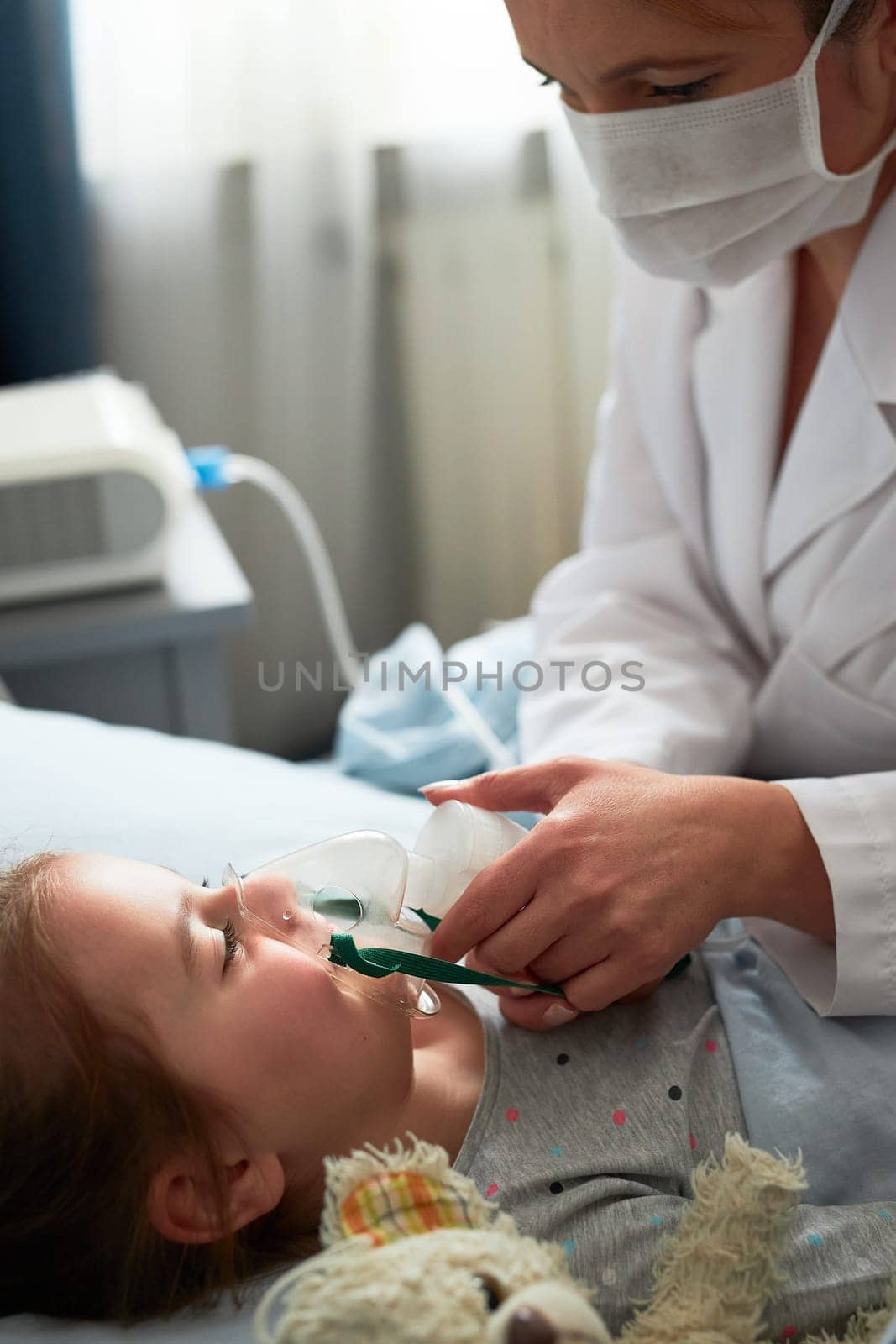 Doctor visiting little patient at home. Child having medical inhalation treatment with nebuliser. Girl with breathing mask on her face. Woman wearing uniform and face mask