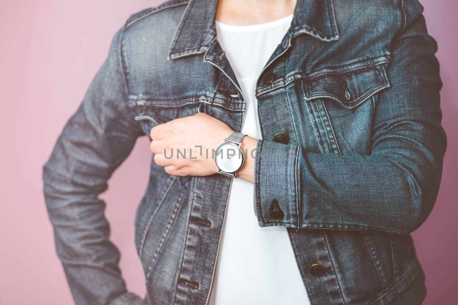 Woman wearing silver wristwatch and jeans jacket. Standing in front of pastel pink wall