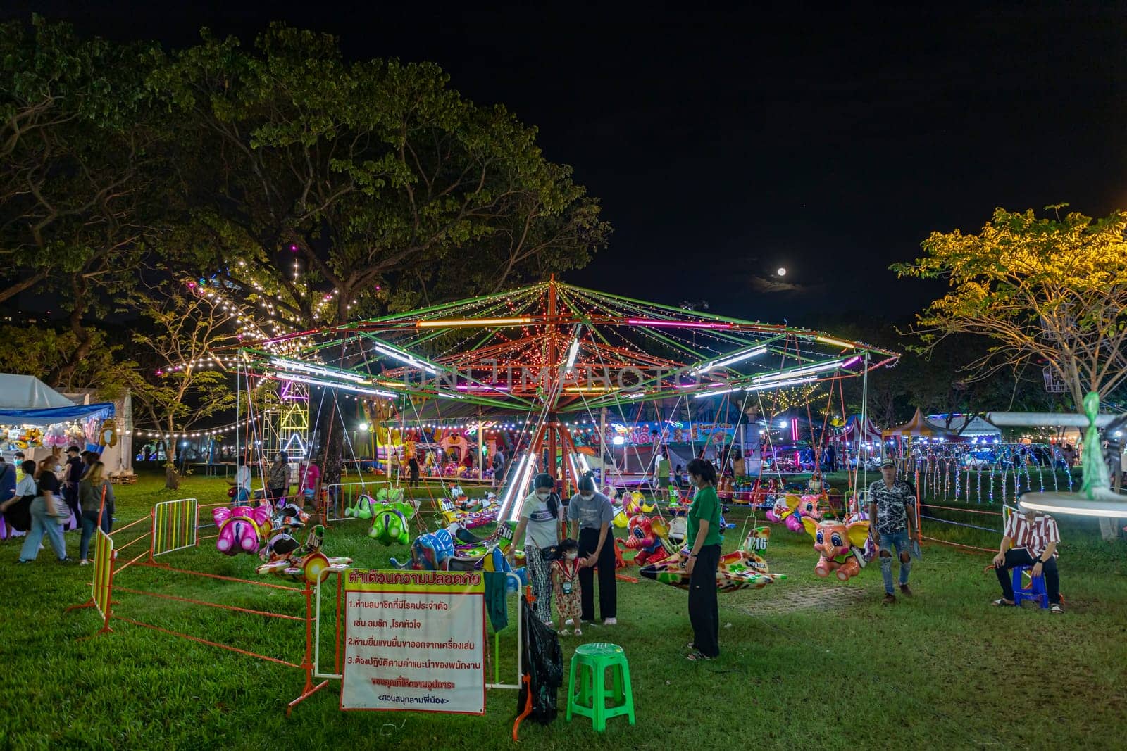 Bangkok, Thailand - Dec 8, 2022 : Crowds of people walk into a beautifully lit amusement park during the Red Cross Fair to be held from December 8-18, 2022. The image is blurred by the movement.