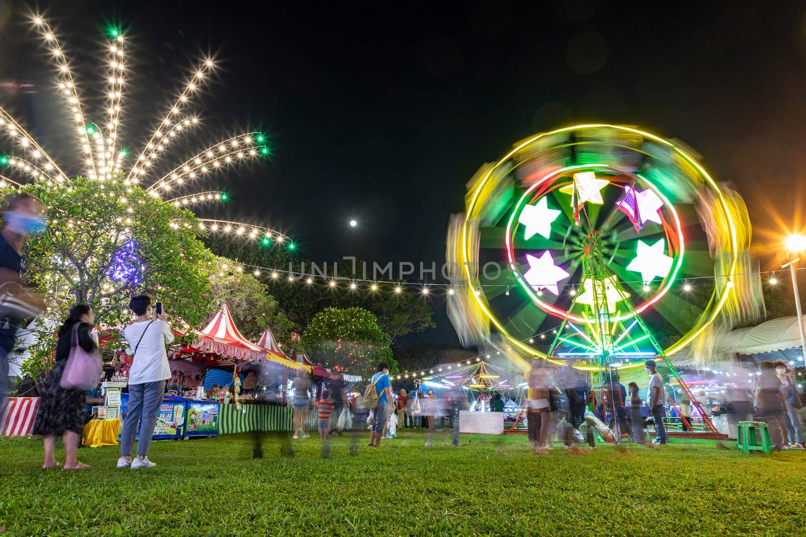 Bangkok, Thailand - Dec 8, 2022 : People take a walk near the spinning Ferris wheel at the Red Cross Fair at Lumpini Park from December 8-18, 2022. The picture is blurred by movement.