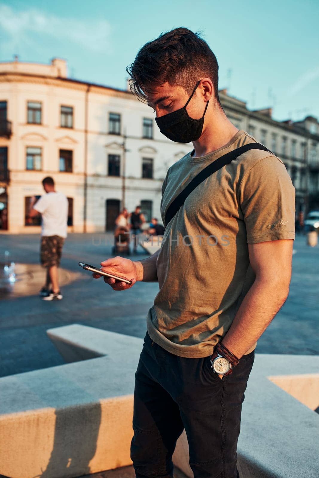Young man standing in the city center in the evening, using smartphone, wearing the face mask to avoid virus infection and to prevent the spread of disease in time of coronavirus