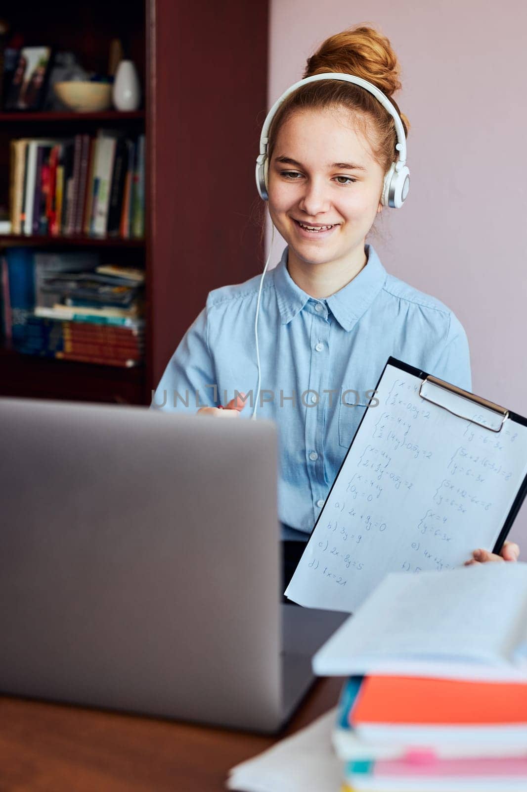 Young woman student showing homework, having classes, learning online, watching lesson remotely, listening to professor, talking with classmates on video call from home during quarantine. Young girl using laptop, headphones, books, manuals sitting at a desk