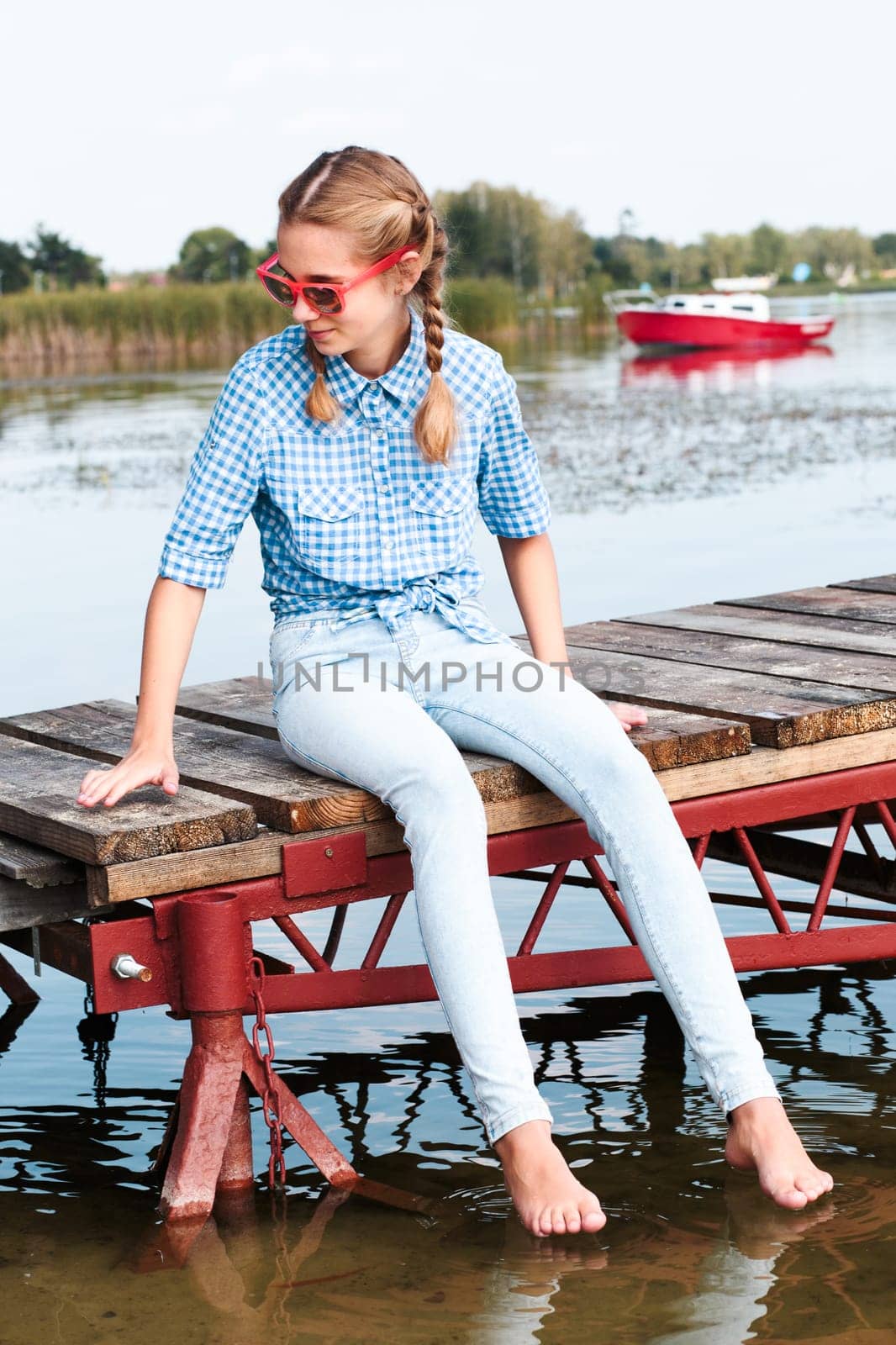 Young girl sitting on jetty over the lake and dipping feet in water on sunny day in the summertime